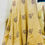 Thumbnail: A close-up view of a high-waisted, flared linen skirt displayed on a mannequin. The skirt is mustard yellow and features intricate hand block printed patterns in maroon, including swirling motifs and stylized floral elements. The fabric drapes gracefully to the floor, emphasizing the fullness and flow of the design. A mirror in the background partially reflects the back of the mannequin, showing the texture and craftsmanship of the skirt from another angle