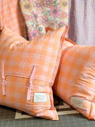 Back view of square envelope cushion with side flange and ties and rectangle cushion with flange both in melon and pink gingham seersucker fabric sitting on workroom table