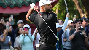 Golfista realizando un swing rodeado de espectadores con móviles. Fondo de edificio rojo y verde. Camiseta negra y gorra blanca. Ambiente expectante.