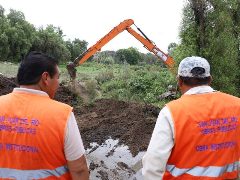 Dos obreros con chalecos naranjas observan una excavadora naranja excavando tierra en un campo; texto: San Juan del Río.