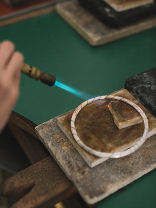 יד אוחזת מבער דולק ומחממת חוט כסף שנמצא על לבנה התלמה על שולחן הצורפות