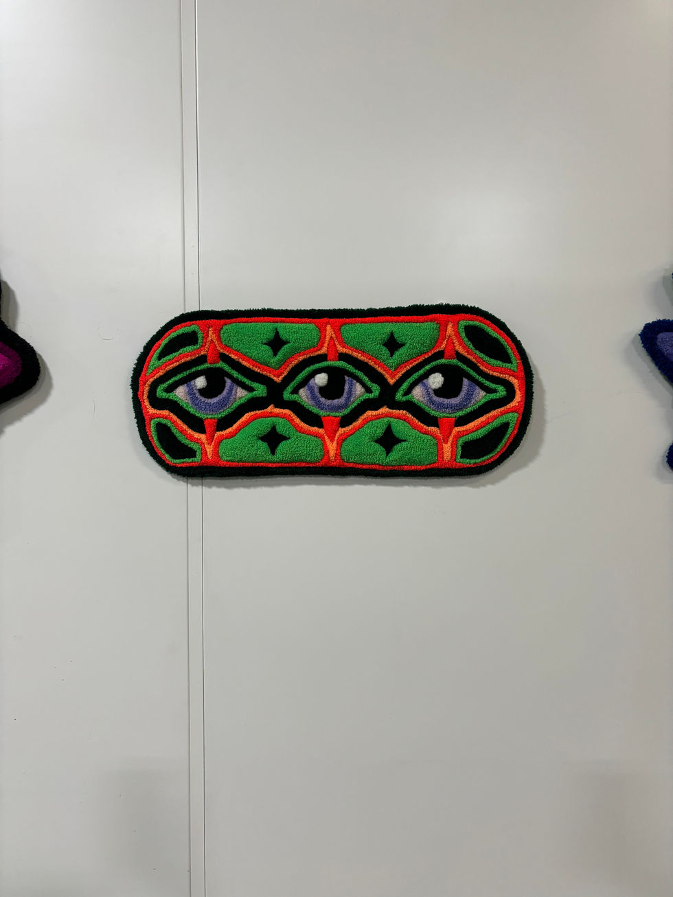Oval shaped tufted rug with three blue-colored eyes lined up in a row. The eyes are outlined with two shades of green and two shades of orange. There are diamond motifs in the surrounding area.