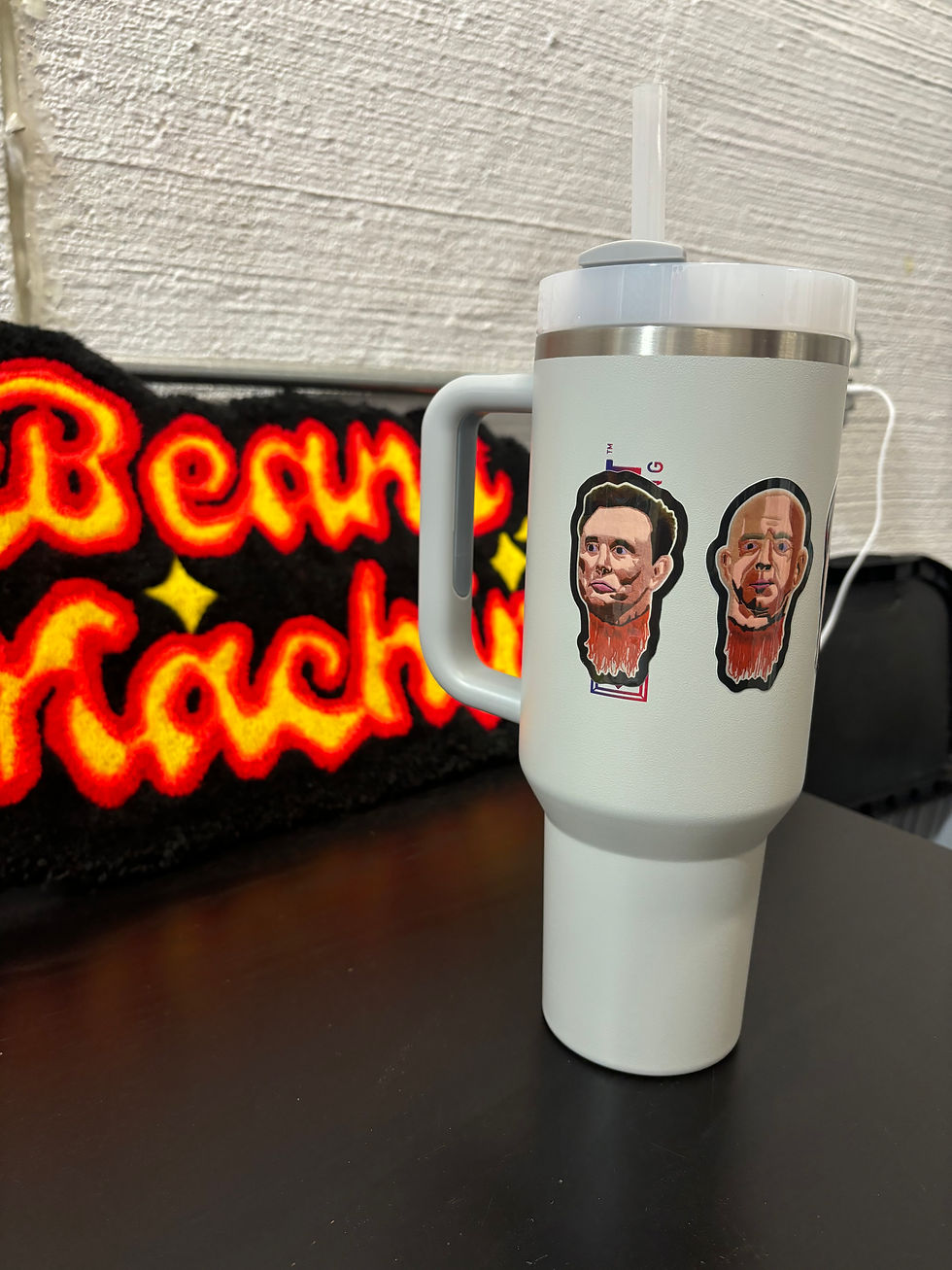 Image of the Musk and Bezos sticker on a gray water bottle. In the background is a rug that says "Beans Machine".
