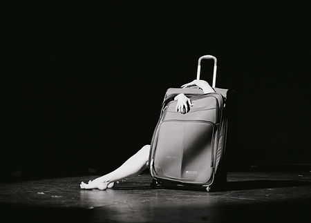 black and white photo of dancer sitting inside of an upright suitcase, her legs outside of it