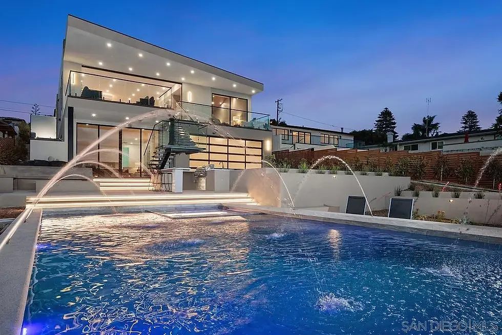 Modern house with glass walls and a lit interior overlooks a blue pool with water jets. Evening sky adds a serene atmosphere.