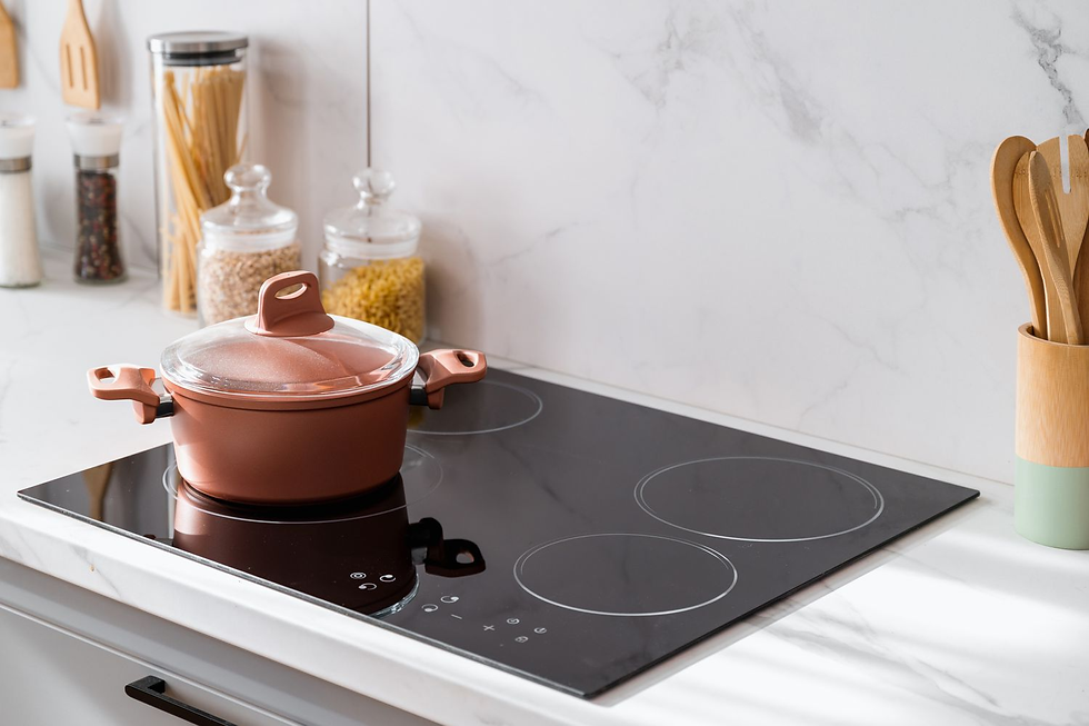 A brown pot with a lid sits on a black induction stove. Kitchen contains jars of pasta, spices, and wooden utensils on a marble counter.