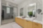 A smart mirror bathroom. A large, frameless glass shower enclosure, fitted with a rain shower head, is situated on the left side of the image. The shower walls are clad in gray rectangular tiles, and the shower floor is composed of small, speckled stones. A vanity with a light-colored wooden cabinet is positioned to the right of the shower. The vanity top is a pristine white quartz or marble, which is also visible in the sink basin. The cabinets have dark-colored, simple, modern handles or pulls. The wall behind the vanity is covered with a striking mosaic tile pattern in shades of gray and white, with a design resembling blooming flowers or similar abstract shapes. A large framed mirror is mounted on the wall above the vanity, reflecting the design of the mosaic tile and part of the shower. A smaller, angled mirror is mounted on the wall above the larger mirror. A potted plant sits in the corner of the back portion of the bathroom between the mirror and the shower, partially visible through the mirror. The flooring throughout the bathroom is a light beige or cream-colored, smooth tile or laminate. A white bath mat or towel is placed near the shower opening. A light is visible above the glass shower door. The overall impression is one of a sophisticated and contemporary bathroom.