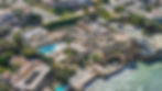 Aerial view of a grand seaside mansion with a pool, ornate architecture, and lush gardens. Waves crash against the cliffside below.