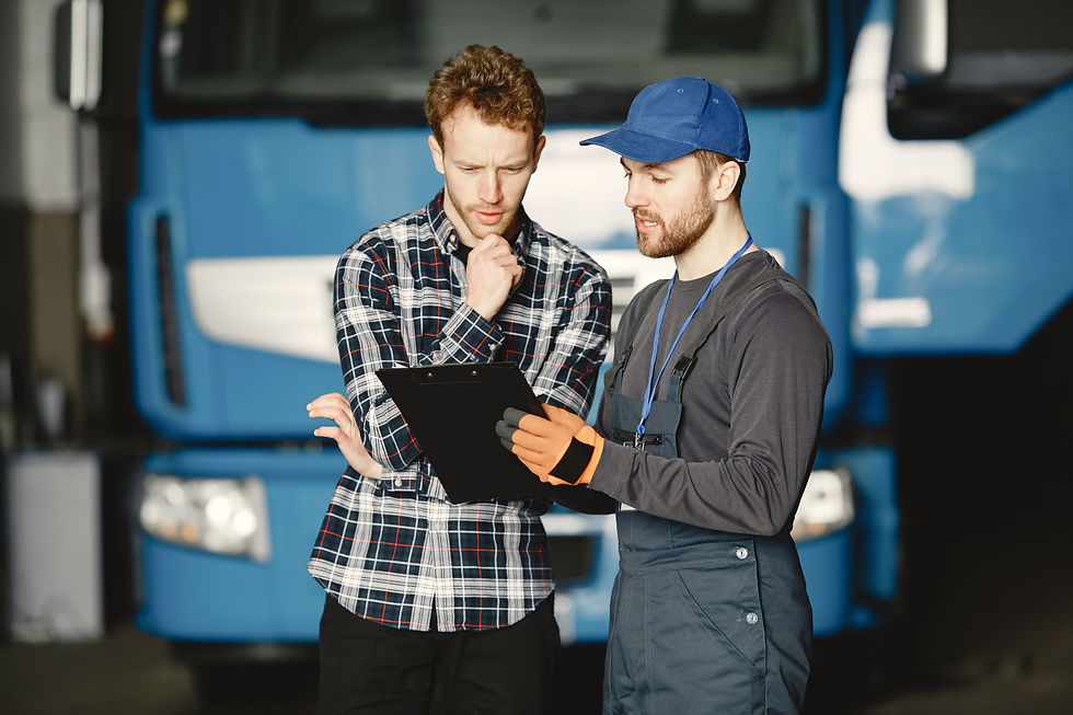 Professional mechanic in blue overalls and cap explains details from a clipboard to a thoughtful owner-operator wearing a plaid shirt. A large blue semi-truck is parked inside the maintenance garage behind them. This interaction represents reliable professional services and support for US trucking businesses, found through Trucking Advisor.