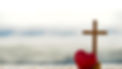 Red heart with wooden Christian cross on gravel floor in morning light, beach sea as backg