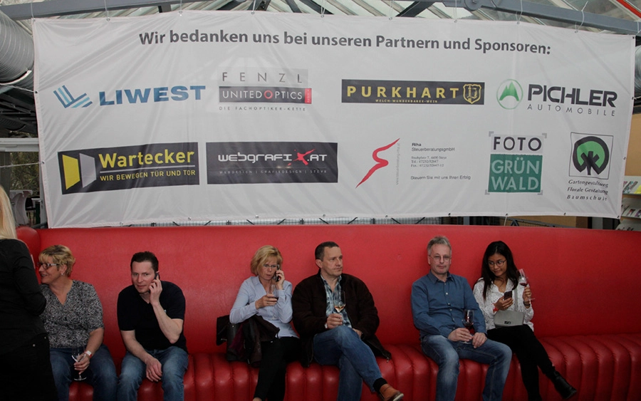 Transparent mit verschiedenen Sponsorenlogos. Davor sitzen mehrere Gäste auf einer roten Bank.