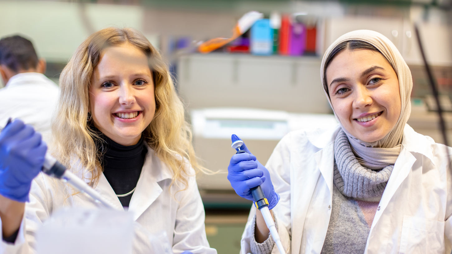 Two women in lab coats smiling while conducting research. The Tam Lab.