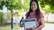 Smiling nurse in pink holds DAISY Award plaque and trophy outdoors with leafy green background.