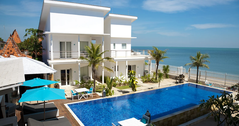 White beachfront villa with palm trees, blue umbrellas, and a sparkling pool. Ocean view in the background under a bright blue sky.