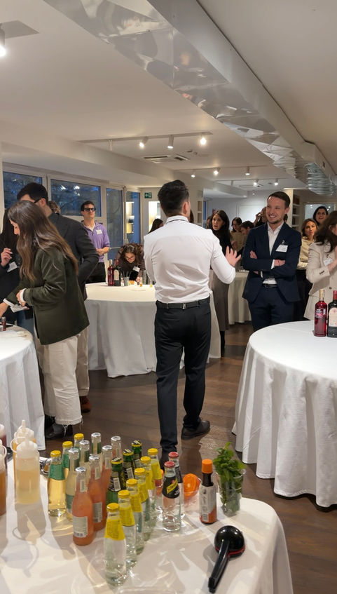 Un bartender formatore, visto di spalle in camicia bianca, illustra le fasi del workshop di mixology a un gruppo di dipendenti aziendali disposti intorno a tavoli circolari con tovaglie bianche. L'atmosfera è professionale e conviviale, con i partecipanti che ascoltano con interesse in una location moderna e luminosa a Milano. In primo piano, un banco bar allestito con un'ampia varietà di soft drink, succhi, guarnizioni di menta e arancia pronti per la sessione pratica.