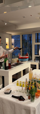 Un bartender professionista in divisa elegante (camicia bianca e farfallino) mostra la preparazione di diversi cocktail colorati durante un workshop aziendale. In primo piano, una fila di drink guarniti con frutta fresca e menta su un tavolo bianco, mentre i partecipanti osservano e scattano foto in una location moderna a Milano.