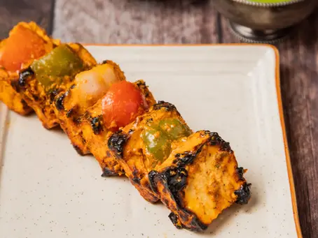 A close-up photograph of grilled paneer tikka skewers arranged on a white plate. Each skewer alternates golden-brown paneer cubes with red and green bell peppers and red onion pieces, all lightly charred. A small bowl of mint sits to the right, two lemon wedges and pickled onion to the left. The lighting is natural, highlighting the warm colors and textures against a clean white plate.