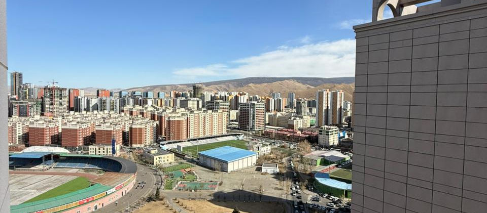 A panoramic aerial view of a vibrant city in Ulaanbaatar, Mongolia, under a clear blue sky. In the foreground, a large stadium with a green field and a running track is visible, surrounded by pathways and dry grassy areas. A busy road with vehicles runs alongside, leading towards a cluster of residential and commercial buildings in the midground. In the distance, numerous multi-story buildings of varying colors fill the cityscape, backed by low-lying hills or mountains on the horizon. The scene captures an urban landscape with recreational facilities and a developing skyline.