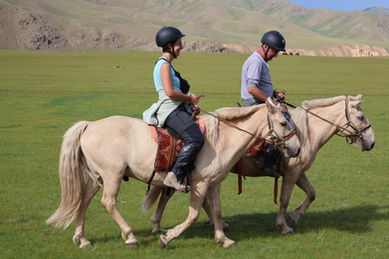 Two individuals riding horses across a grassy, hilly terrain in rural Mongolia. Horse riding, equestrian tour, Mongolian countryside.