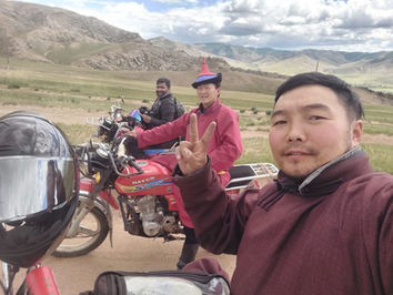 Two people posing with motorcycles, one looking at the camera, in a scenic outdoor setting in Mongolia. Motorcycle travel, friendly locals.