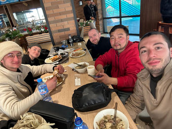 Group of people gathered around a table eating and socializing in a warm, rustic indoor setting, possibly a guesthouse in Mongolia. Travel group, mealtime, camaraderie.