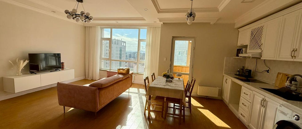A spacious, sunlit open-concept living area with light wooden flooring. A light pink sofa is centrally placed, facing a large window that offers a view of city buildings. To the left, there's a white TV console with a television. On the right side of the room, a modern kitchen area features white cabinets, a sink, and a washing machine, along with a wooden dining table and chairs. The room is well-lit by natural light streaming through the window and modern light fixtures on the ceiling, suggesting a bright and inviting living space.