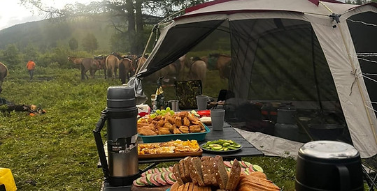 A close-up shot of a camping table laden with a variety of food, including sandwiches, bread, and pastries. In the misty background, a tent is set up, and a herd of cattle is visible grazing on the hillside.