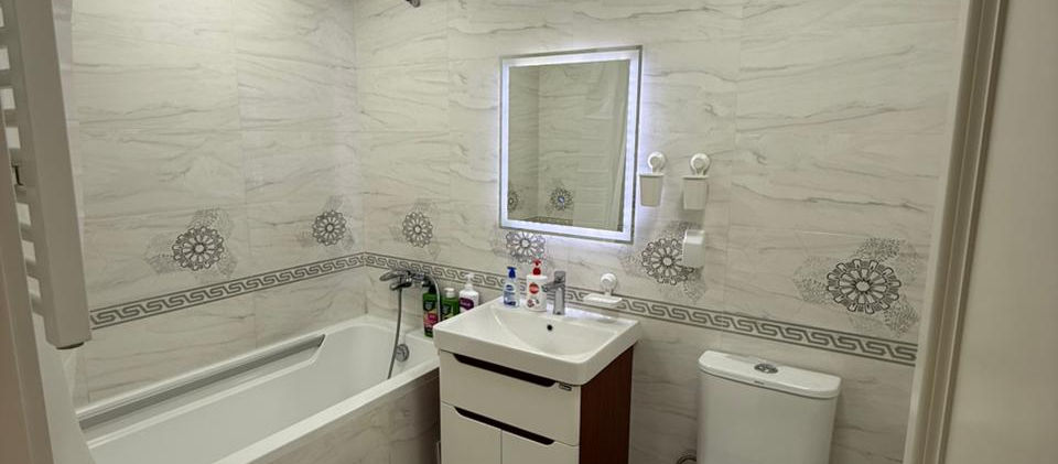 A clean, modern bathroom with white marble-patterned tiles on the walls and floor. It features a white bathtub on the left, a vanity with a white sink and a brown cabinet, and a toilet on the right. Above the vanity, there's a rectangular mirror with integrated lighting. The room is well-lit by overhead square ceiling lights and has decorative border tiles and patterns on the walls.