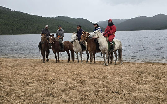 A group of five horseback riders and their horses stand on the sandy shore of a serene lake. The riders are dressed in warm clothing, and the background features rolling, tree-covered hills under a cloudy sky.