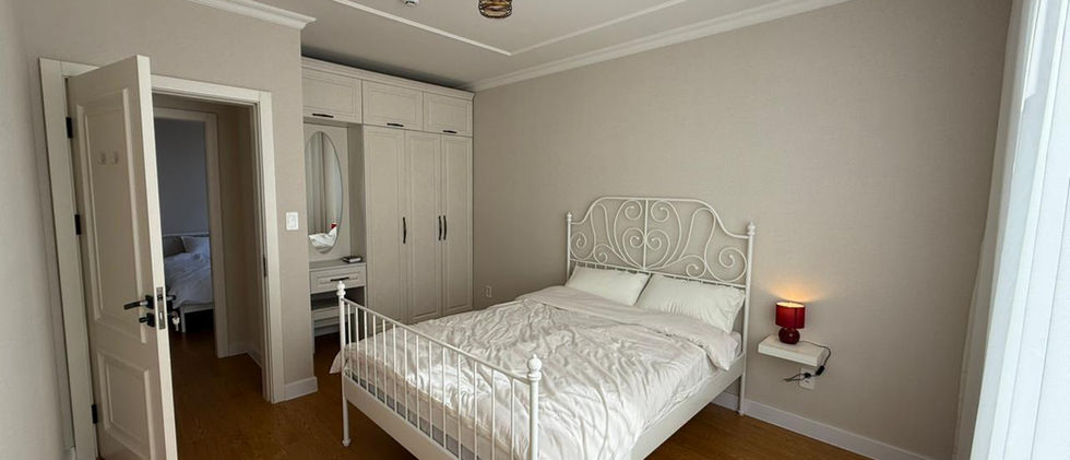A bright bedroom with light wooden flooring and neutral-colored walls. A white metal-framed double bed with a white duvet and pillows is centrally placed. On the right side of the bed, a small floating shelf with a red lamp is mounted on the wall. In the background, there's a light-colored wardrobe with multiple doors and a vanity area with a mirror. A doorway on the left leads to another room, offering a glimpse of a bed. The room is well-lit and has a decorative ceiling.