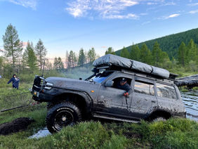 Off-road jeep with rooftop tent driving through a muddy track in a lush green landscape with trees and distant mountains. Off-roading adventure, overland travel, remote exploration.