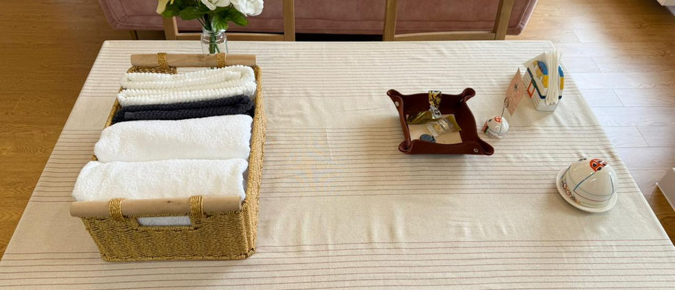 A close-up overhead shot of a wooden dining table covered with a light-colored, striped tablecloth. On the table, there's a woven basket filled with neatly folded white, black, and gray towels, suggesting guest amenities. To the left, a vase holds a bouquet of white and yellow artificial flowers. Other items on the table include a decorative brown dish, a small bell, and a tissue box. In the background, a light pink sofa is visible, indicating the dining area is part of an open living space.