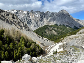 Dramatic mountainous landscape with rocky slopes, scattered pine trees, and snow patches under a cloudy sky. Mountain scenery, rugged terrain, nature photography.