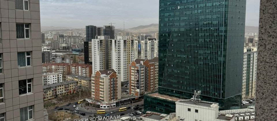 An aerial view of a bustling city center in Ulaanbaatar, Mongolia, seen from a high vantage point. In the foreground, the 'NAADAM CENTER' building is prominent, featuring retail stores and a distinctive blue-roofed section. To its right, a modern, tall green glass building, likely the Khan Bank tower, stands out against the skyline. Surrounding these main structures are various other multi-story residential and commercial buildings, along with a busy street with cars and buses. Distant mountains or hills are visible on the horizon under a partly cloudy sky, indicating the city's geographical setting.