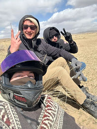 Two people bundled up in warm clothing on a motorcycle, one looking at the camera, in a snowy or cold outdoor environment. Winter motorcycle journey, cold adventure.
