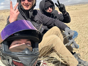 Three people are posing for a selfie while sitting on what appears to be a camel or other large animal in a sandy, desert-like setting. One person is wearing sunglasses and smiling, while the other gives a thumbs-up. The perspective suggests they are on an adventure or touring a desert landscape.