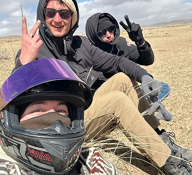 Group of three friends resting on the ground. Two of t he wearing black hooides and black glassess and the last one wearing a motorcycle helmet