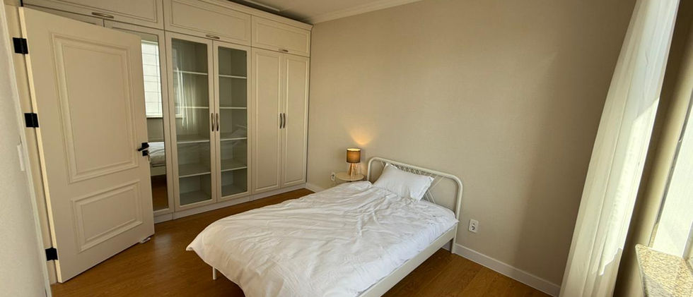 A tidy bedroom featuring light wooden flooring and neutral-colored walls. A single white bed with a white duvet is placed in the center of the room. To the left, there's a large, built-in white wardrobe with some glass-fronted sections and a partially open door, revealing a mirrored surface. A small nightstand with a lamp is next to the bed. On the right, a window with sheer white curtains allows natural light into the room. The room has a simple, clean aesthetic.