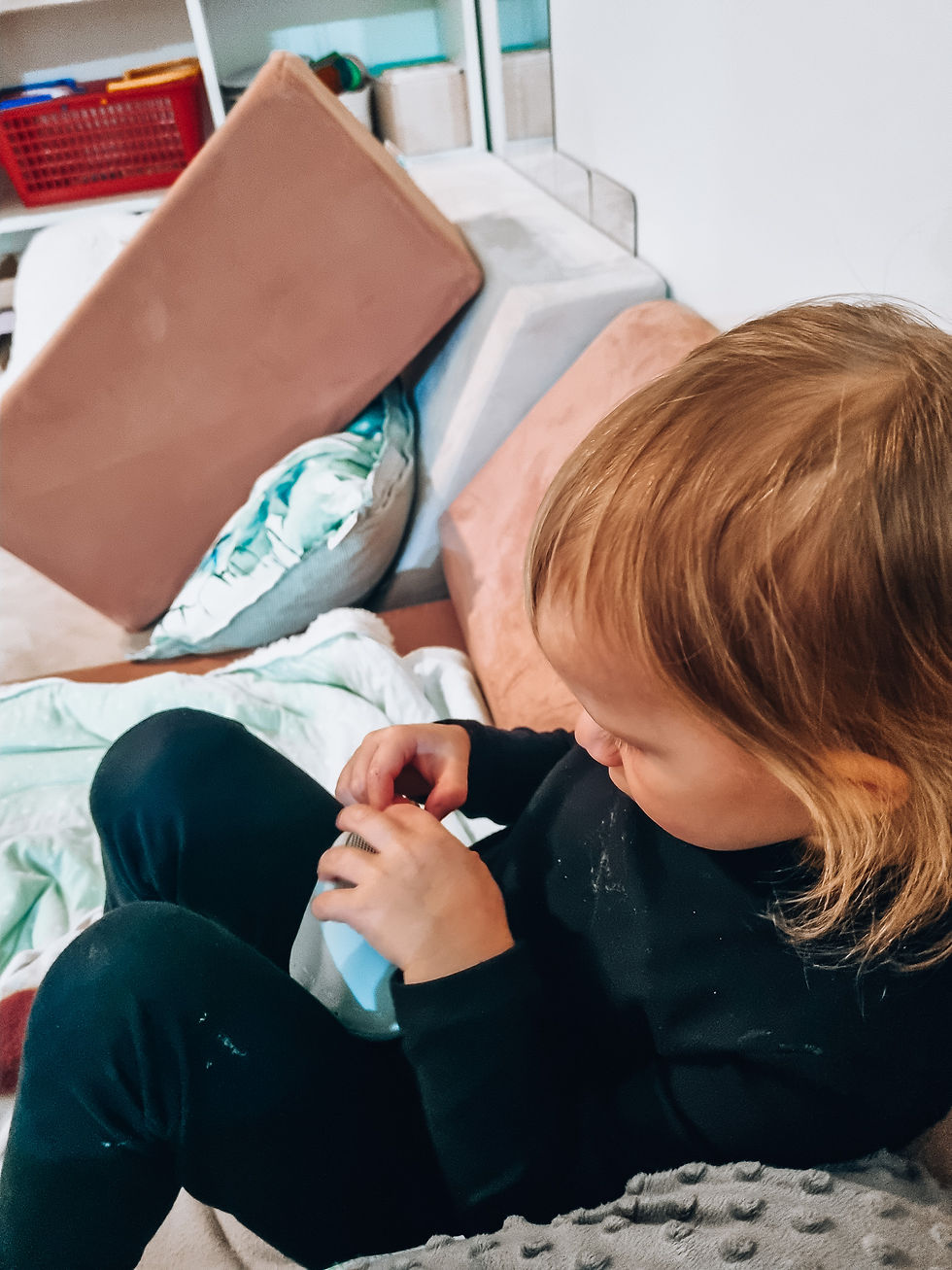 Child in black clothes plays with a cup on a sofa. Background has colorful toys and cushions, creating a cozy, playful atmosphere.