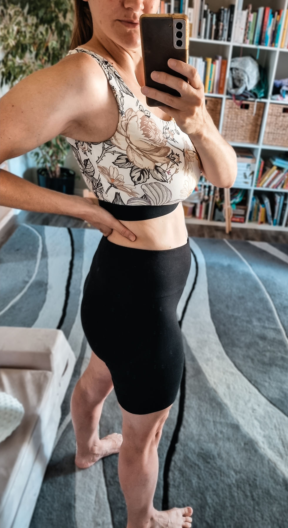 Woman takes a mirror selfie at home in a floral sports bra and black shorts, with bookshelves and a striped rug behind her.