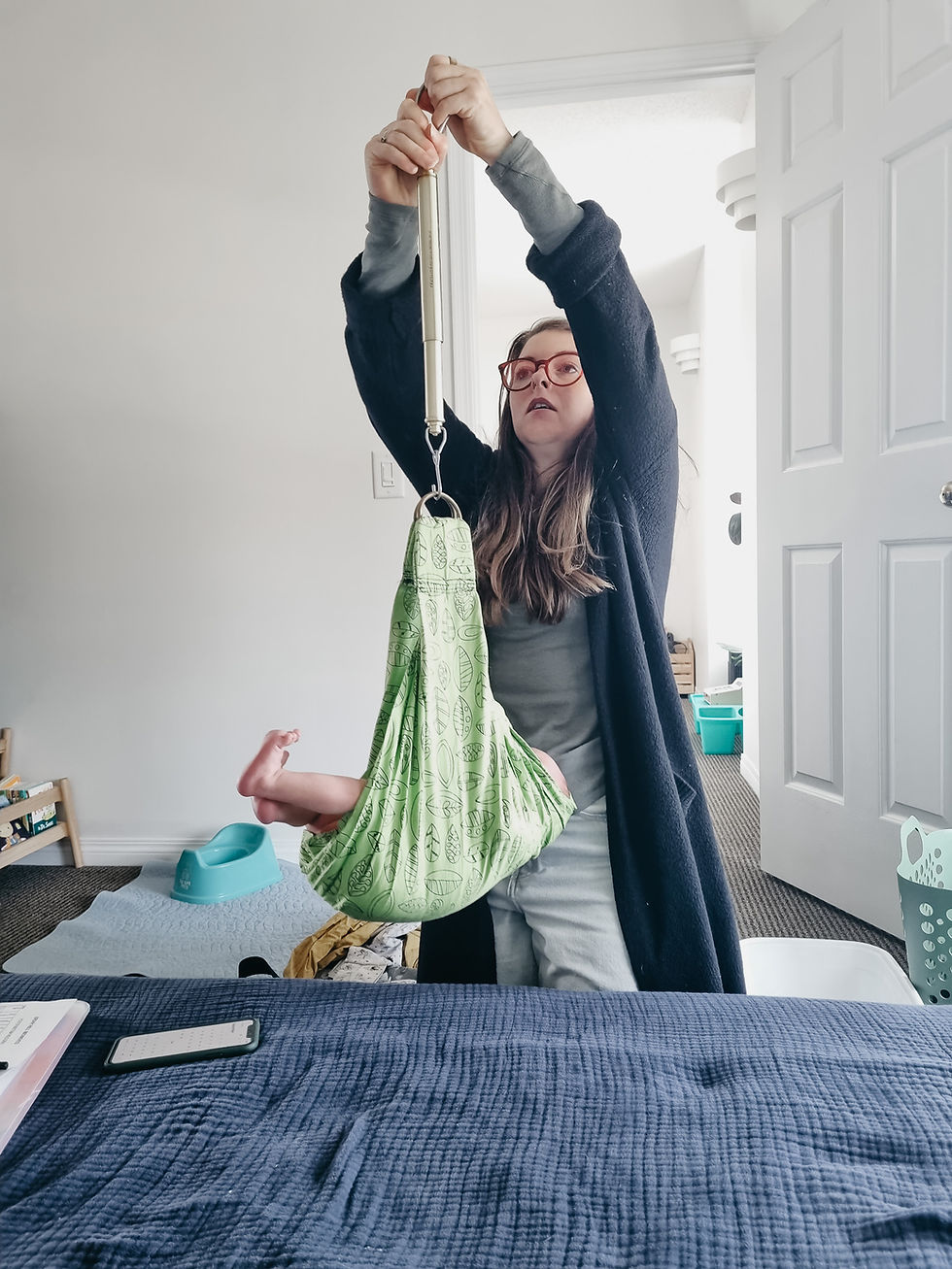A person with red glasses weighs a baby in a green sling in a room with a navy blanket and teal potty chair. They appear focused.
