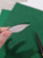 Hand holding a small green felt leaf with a paper pattern on top. Scissors and green felt sheets are in the blurred background.