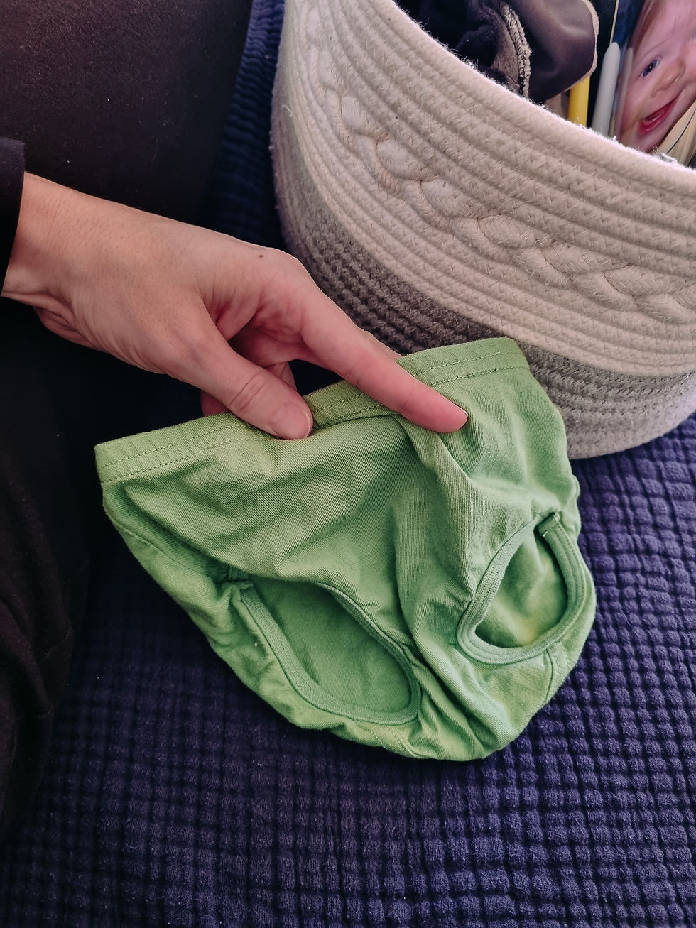Hand holding green toddler underwear on a dark textured surface, next to a white woven basket with a playful, smiling image inside.
