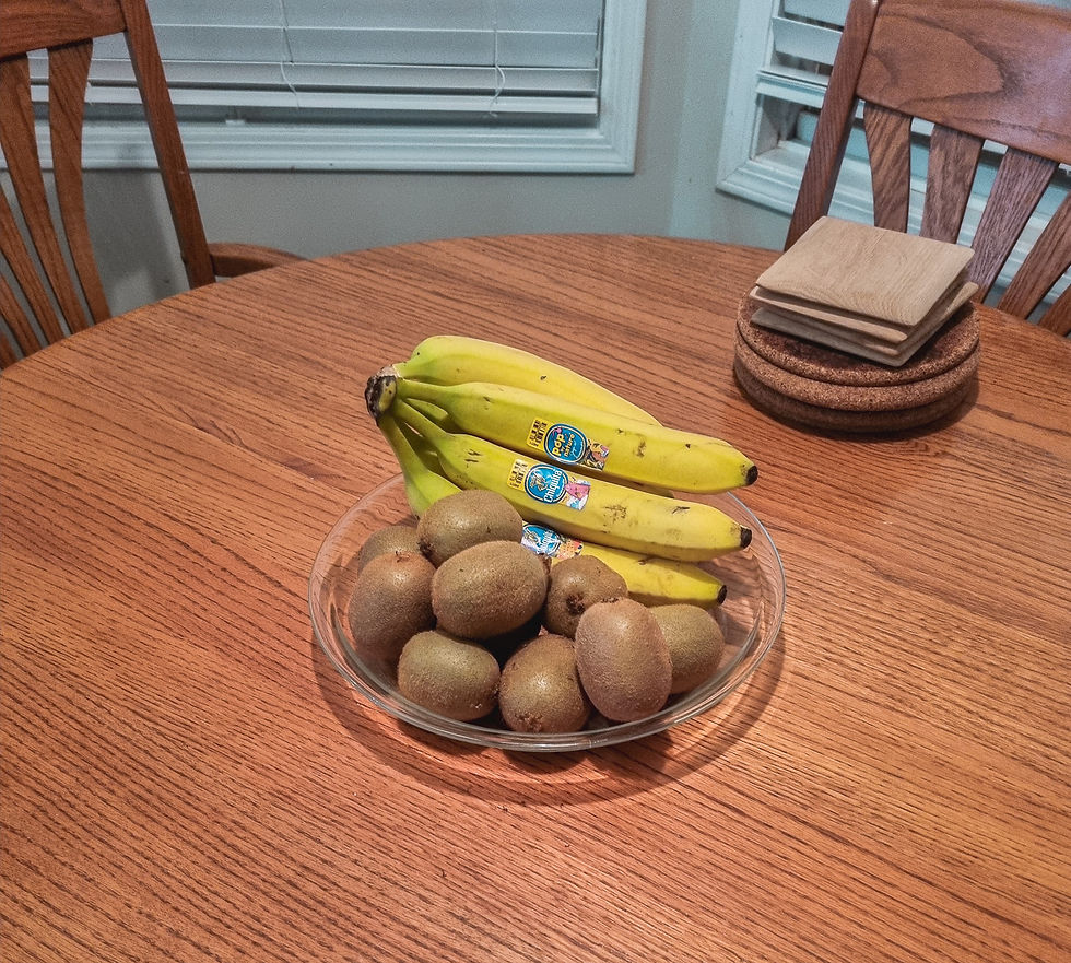 Bowl of kiwis and bananas on a wooden table with chairs, coasters nearby. Labels visible on bananas. Neutral, domestic setting.