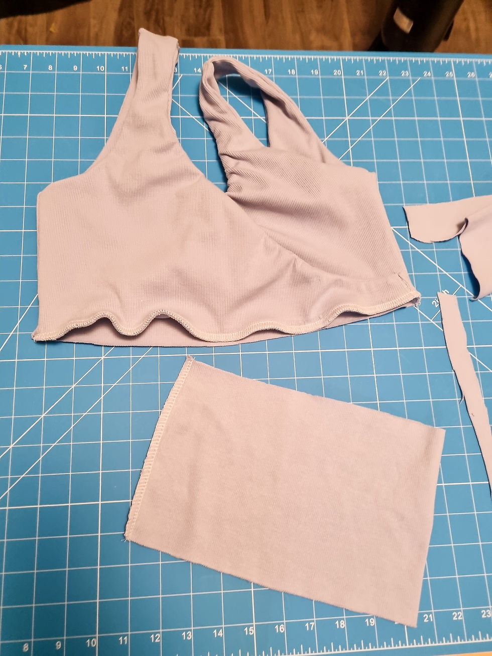 Lilac fabric pieces on a blue grid cutting mat, including a bralette and rectangular piece. Wooden floor background.