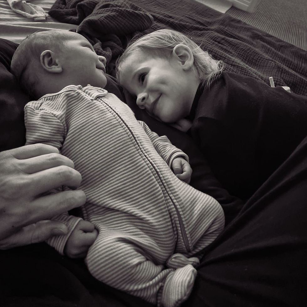 A baby in striped onesie lies beside a smiling older child on a dark blanket. A hand gently touches the baby. Cozy, tender moment.