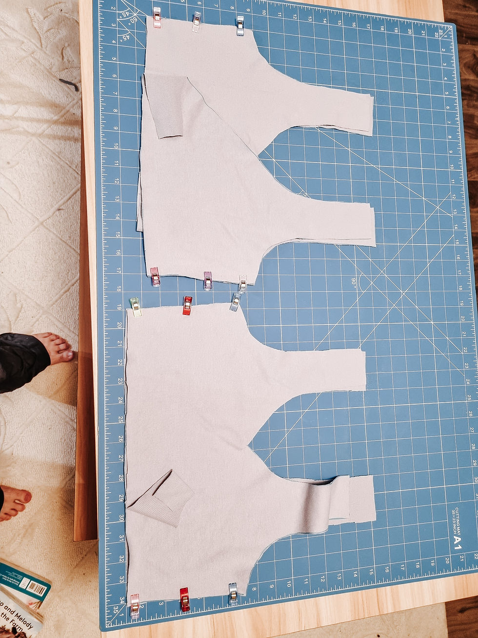 Two gray fabric pieces are clipped on a blue cutting mat with measurement grid. Bare feet visible on the left. Casual indoor setting.