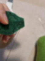 Hand sewing green felt with needle and thread, close-up. A thumb secures the fabric. Background blurred, highlighting the crafting detail.