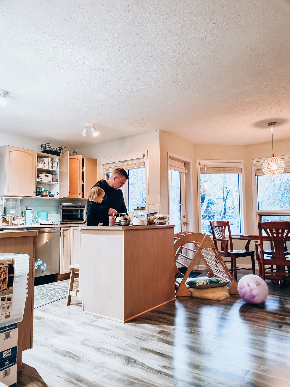 A man and child bake together in a cozy kitchen. Wooden cabinets, scattered toys, and a large window create a warm, inviting atmosphere.