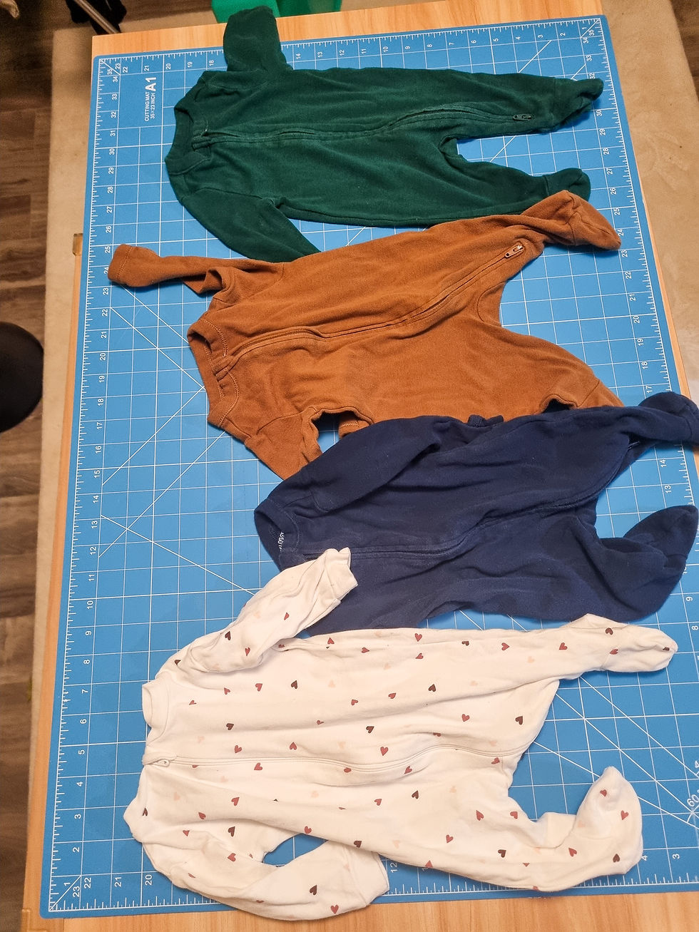 Four baby sleepers in green, brown, navy, and white with hearts are laid on a blue cutting mat with measurements.